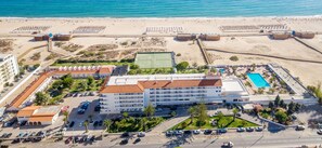 An aerial view of a coastal hotel with a large building, a tennis court, a swimming pool, and a beach area. - Vasco da Gama Hotel (Vila Real Santo Antonio)