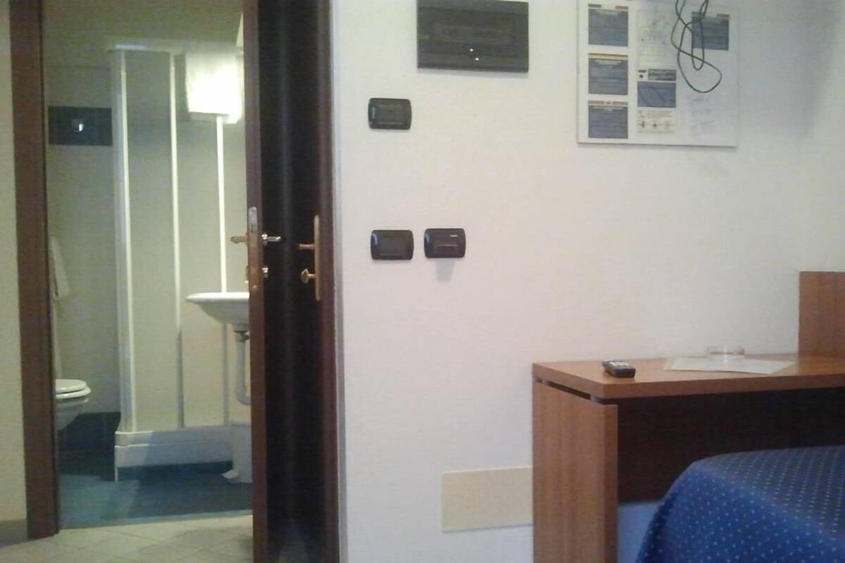 A hotel room with a bed, a desk, a wall-mounted control panel, and a door leading to a bathroom.
