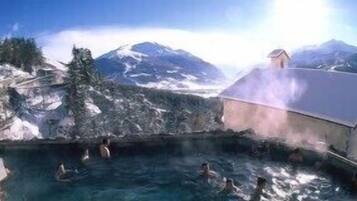 A hot spring with people swimming, surrounded by mountains and a building with a sloped roof.