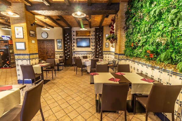 A restaurant with tiled floors, wooden beams, and a green wall with plants. Tables and chairs are neatly arranged. - Hotel Pacoche Murcia (Murcia)