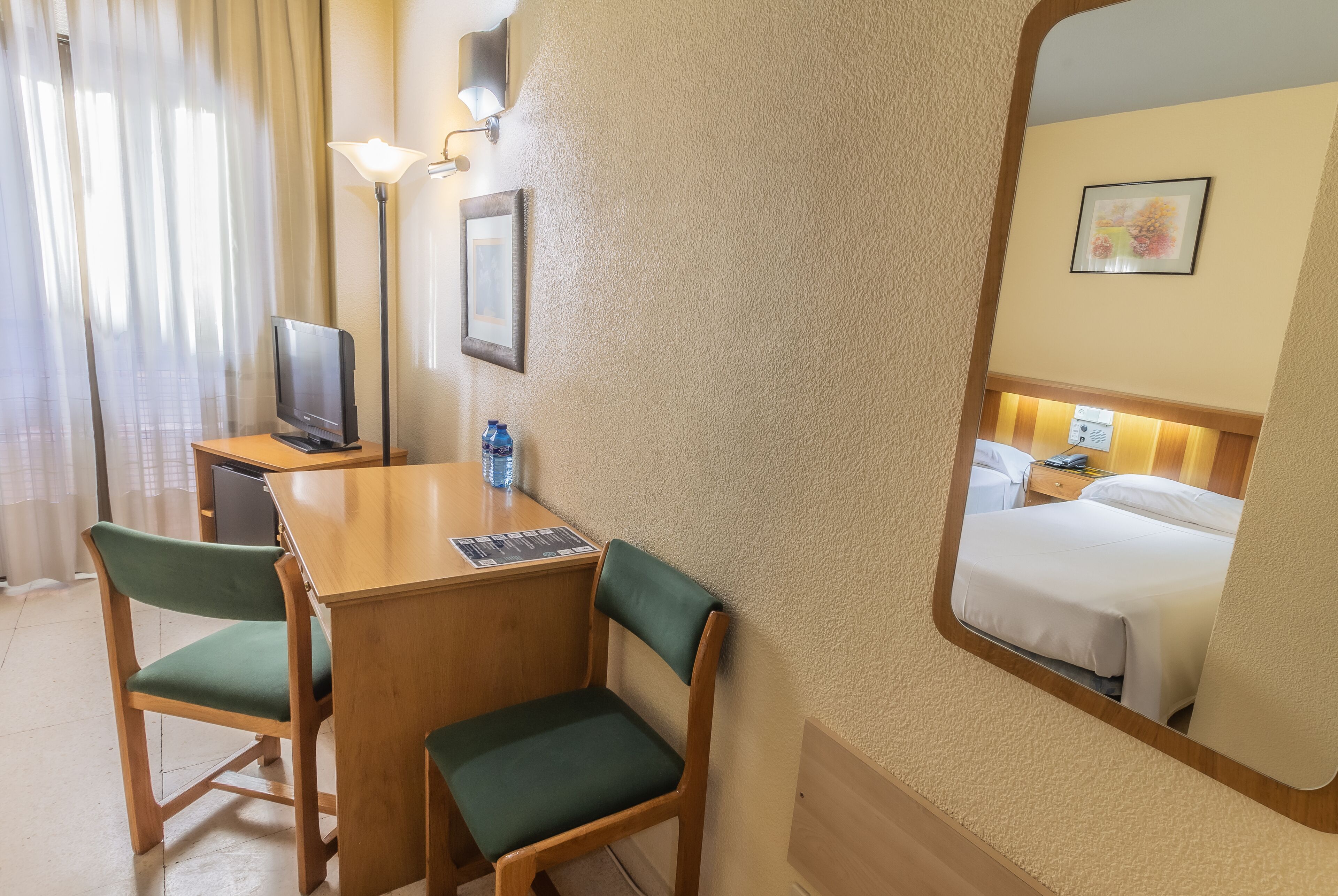 A hotel room with a wooden desk, two green chairs, a television, a water bottle, and a bed with white linens.