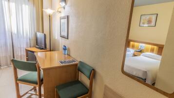 A hotel room with a wooden desk, two green chairs, a television, a water bottle, and a bed with white linens.