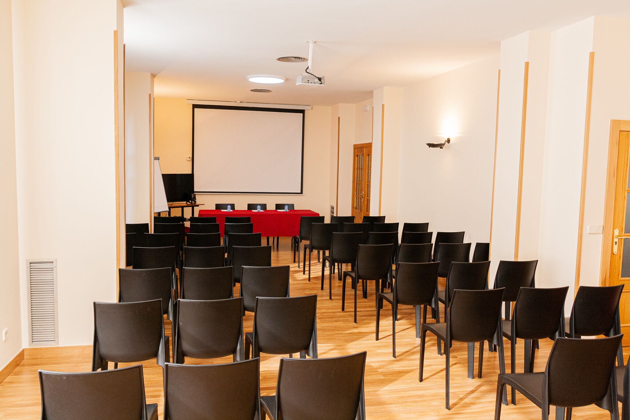 A conference room with rows of black chairs, a red table with a projector screen, and a wooden door.
