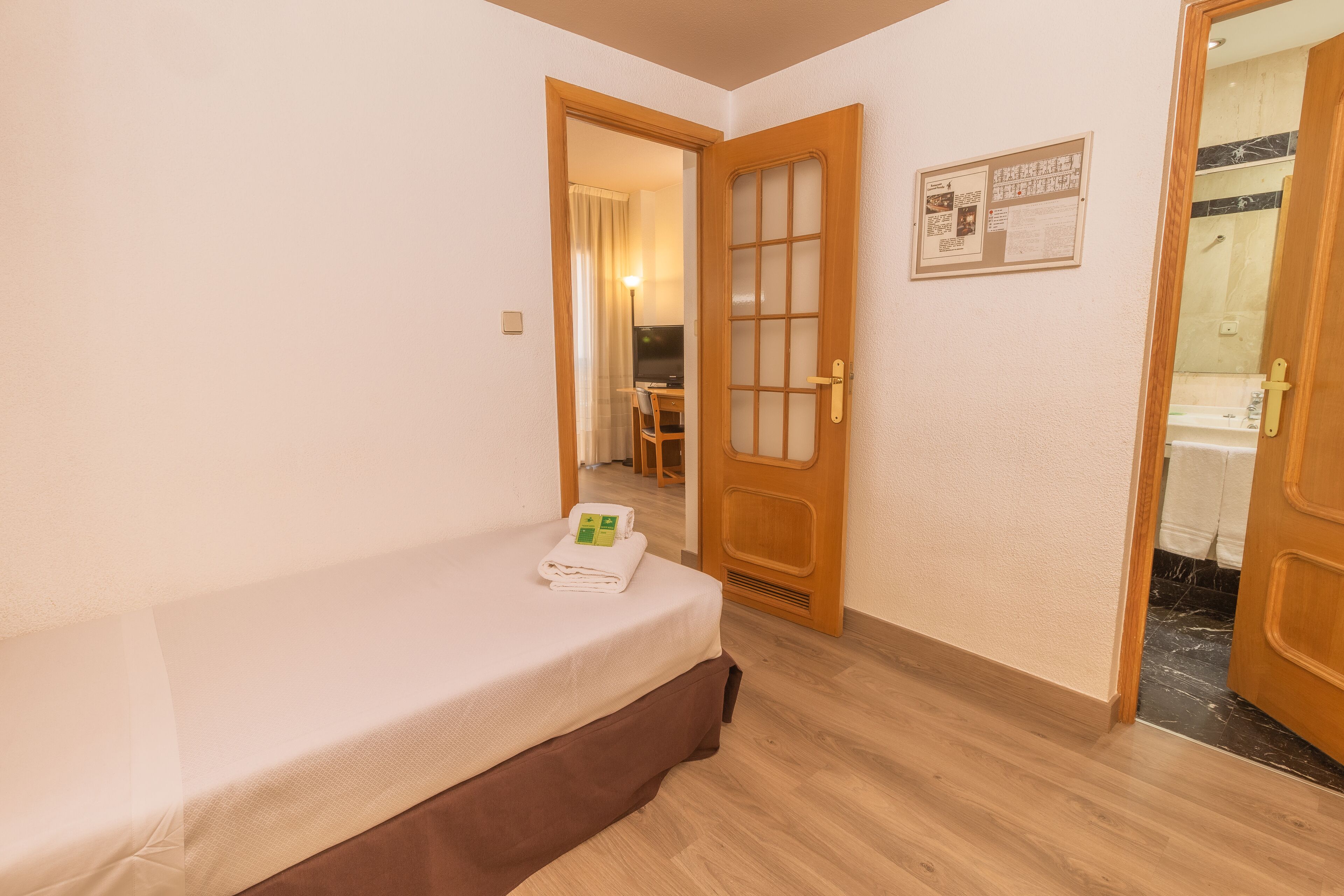Habitación de hotel con cama, suelo de madera, una puerta con panel de vidrio y un baño visible a través de una puerta abierta.