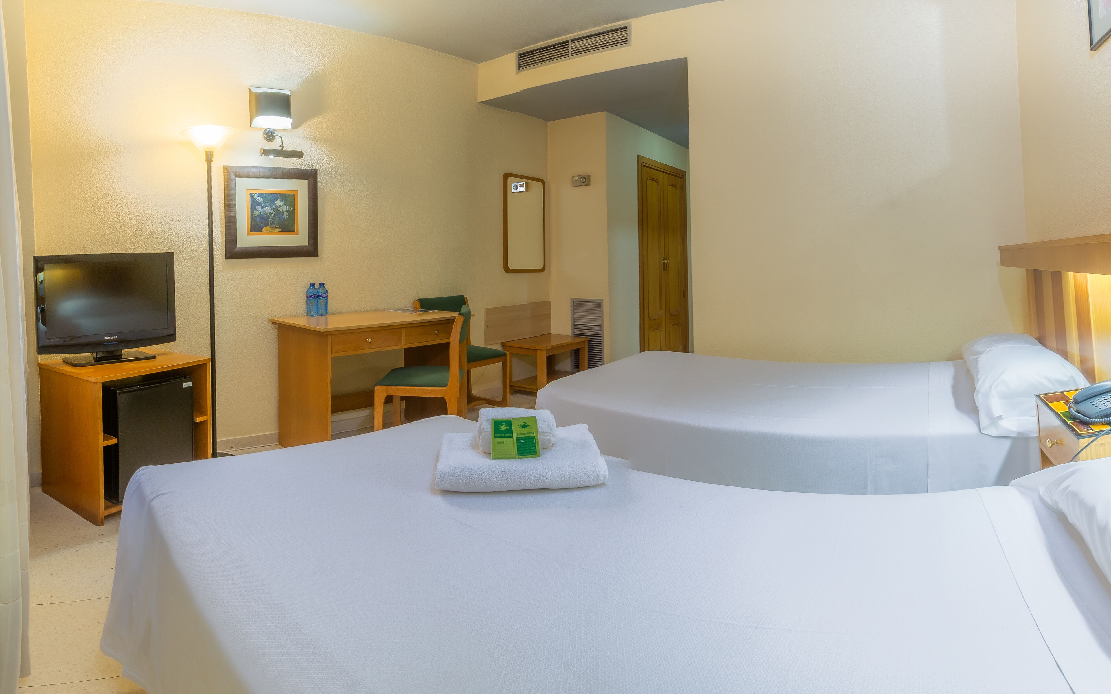 A hotel room with two beds, a desk with a chair, a television, and a small refrigerator.