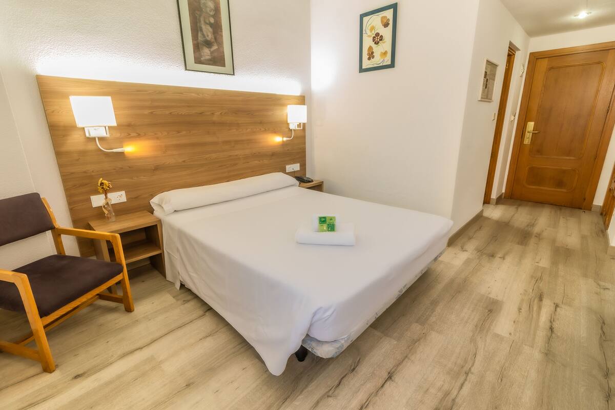 Une chambre d’hôtel avec une tête de lit en bois, un lit simple, une table de chevet avec une lampe, une chaise et une petite table de nuit.
