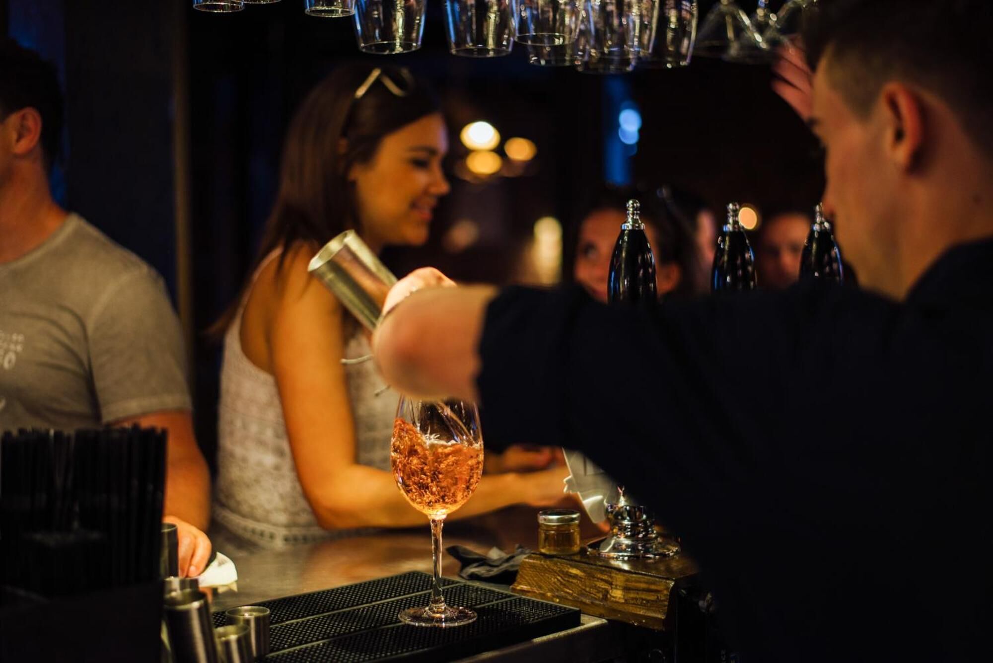 Un barman prépare un cocktail derrière le comptoir, avec différentes bouteilles et un verre à portée de main.