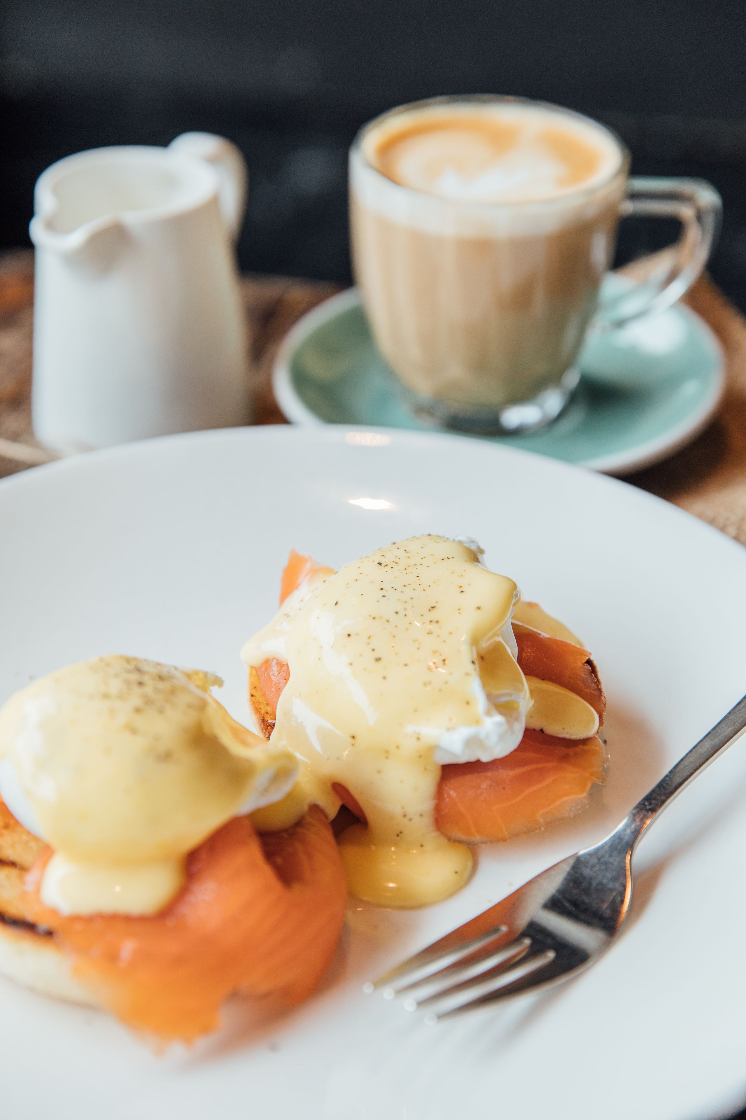 A plate of eggs benedict with salmon and hollandaise sauce, a cup of coffee, and a pitcher of water on a table.