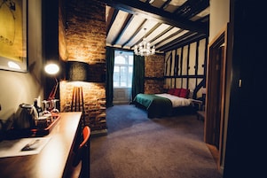 Quarto com paredes de tijolos aparentes, teto de madeira, cama com roupa de cama verde e vermelha, escrivaninha com chaleira e janela com cortinas.