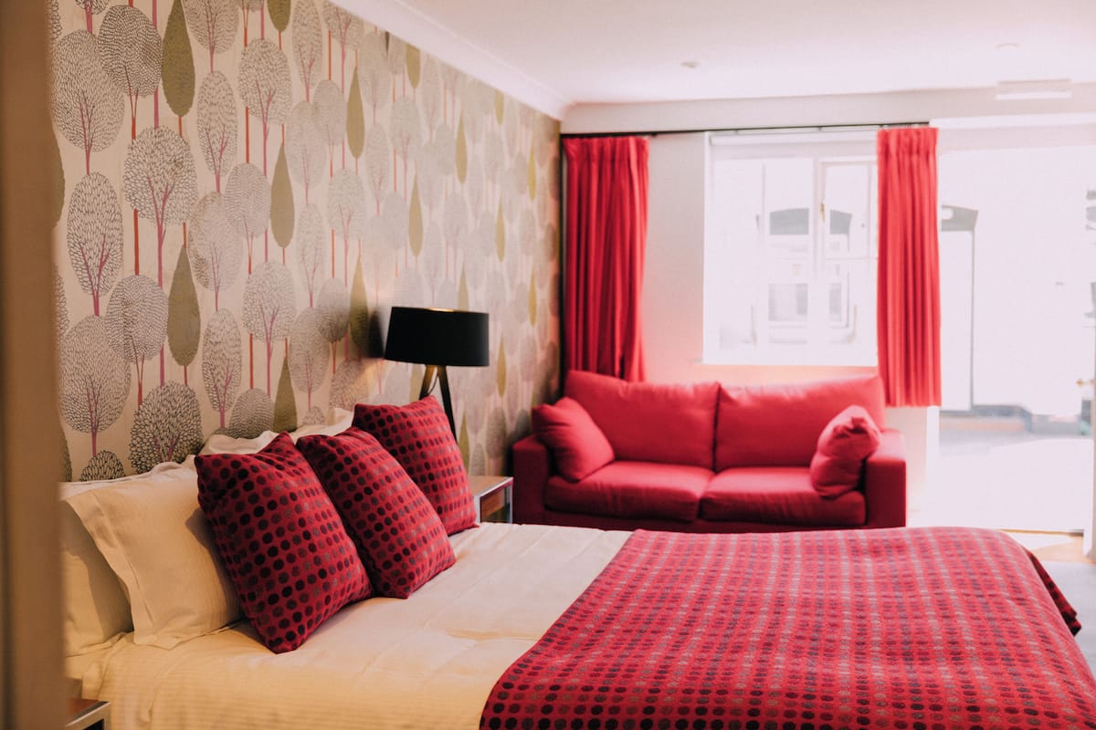 A hotel room with a bed, a red sofa, a nightstand with a lamp, and a window with red curtains.