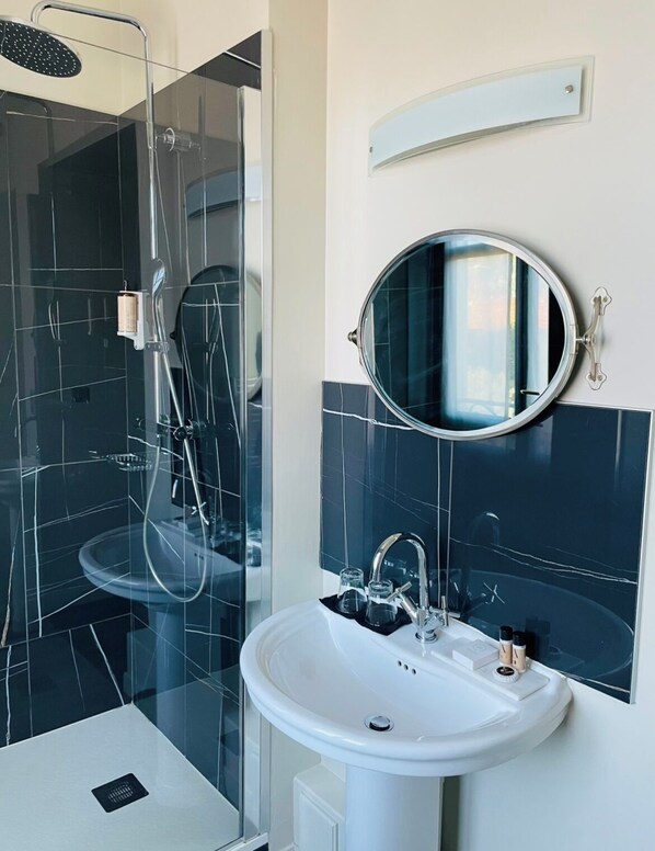 Une salle de bain moderne avec une douche à parois de verre, un lavabo blanc avec un miroir au-dessus, et des murs recouverts de carreaux foncés.
