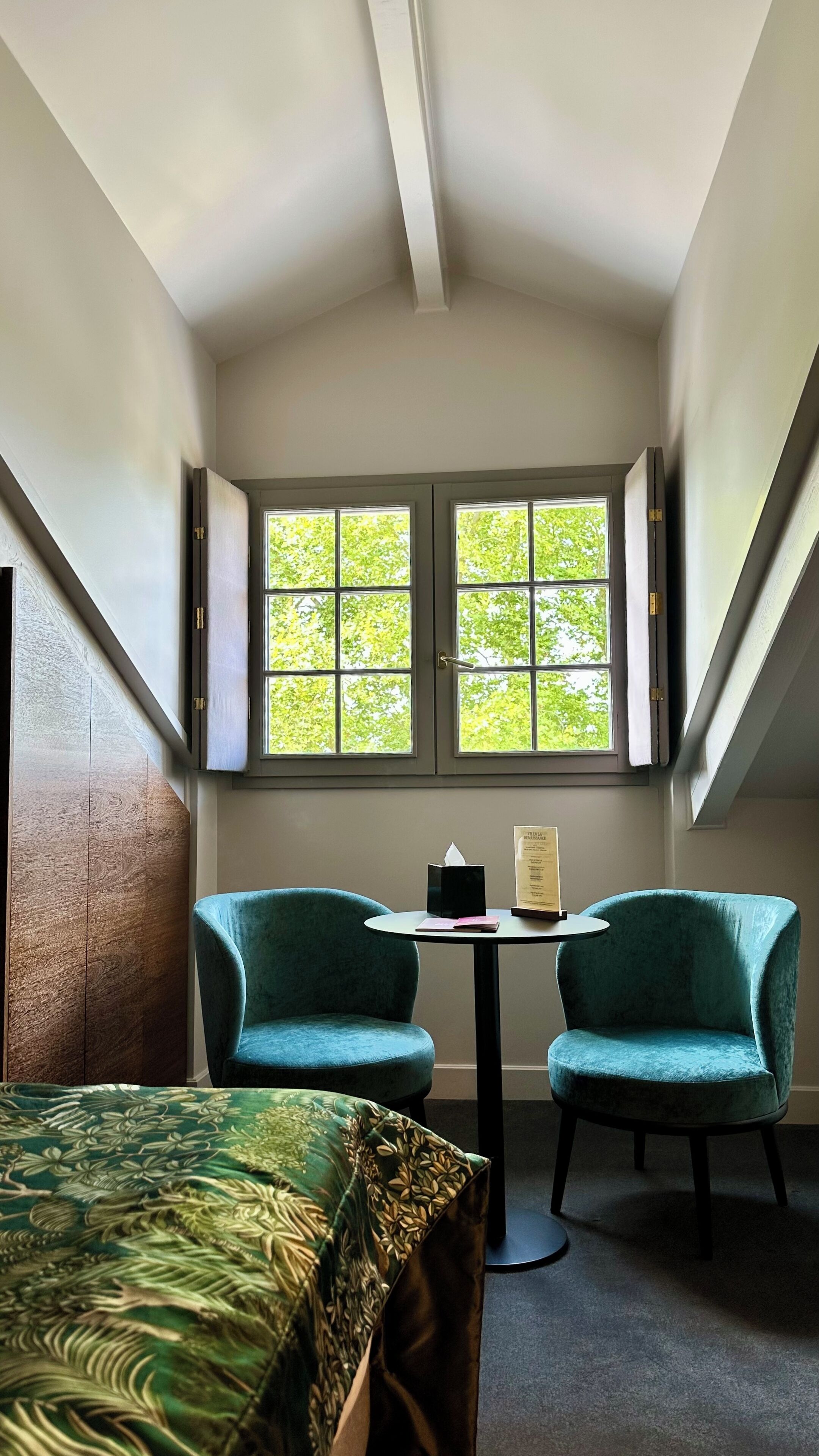 A room with two teal chairs, a small table, and a bed with a green patterned bedspread.
