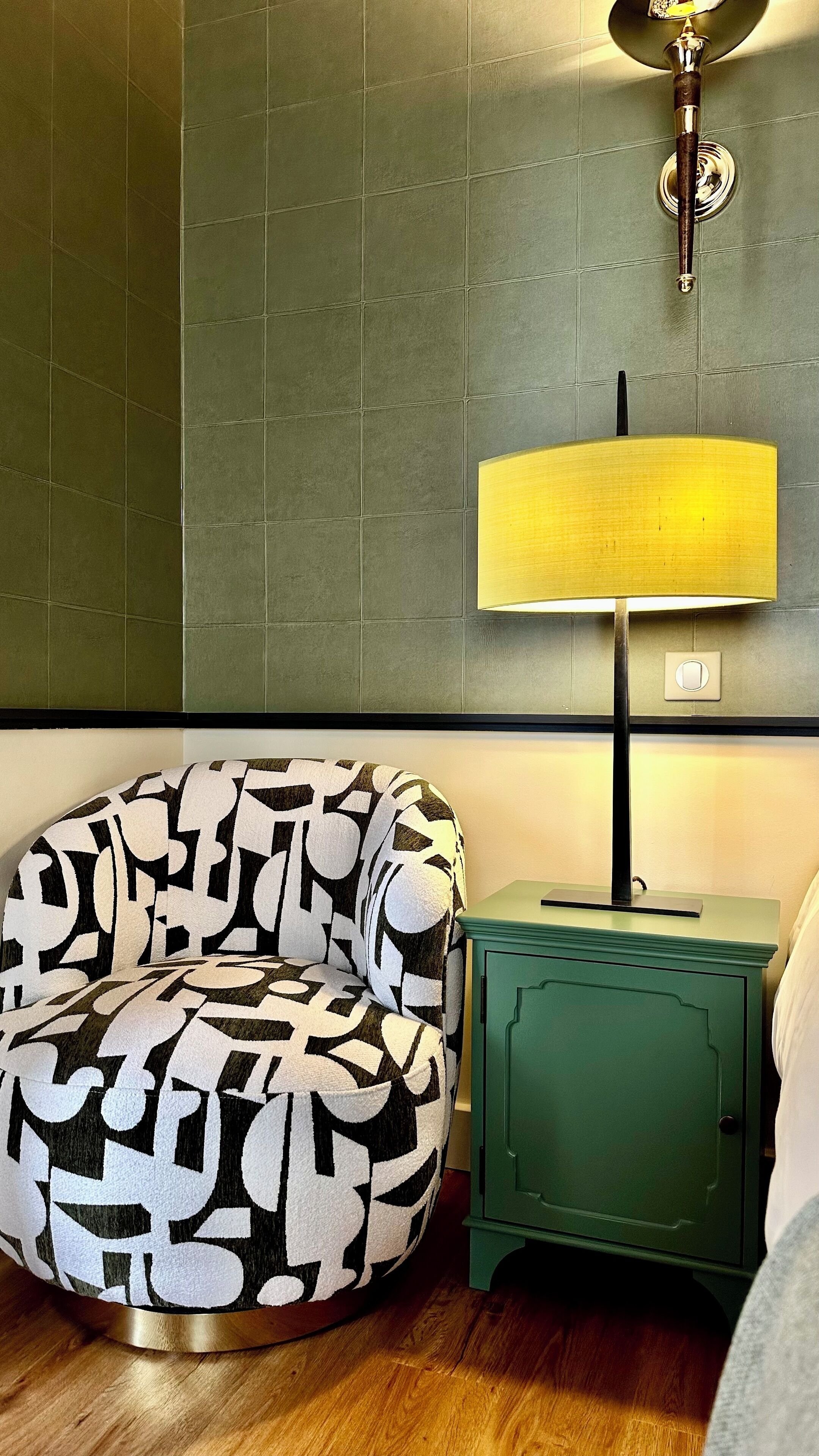 A room with a patterned armchair, a green nightstand, and a wall-mounted lamp.