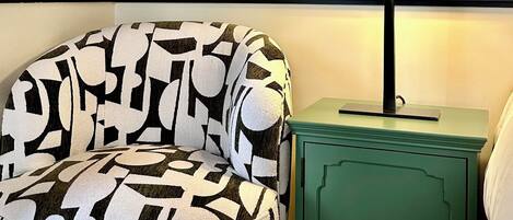 Une pièce avec un fauteuil à motifs, une table de chevet verte et une lampe fixée au mur.