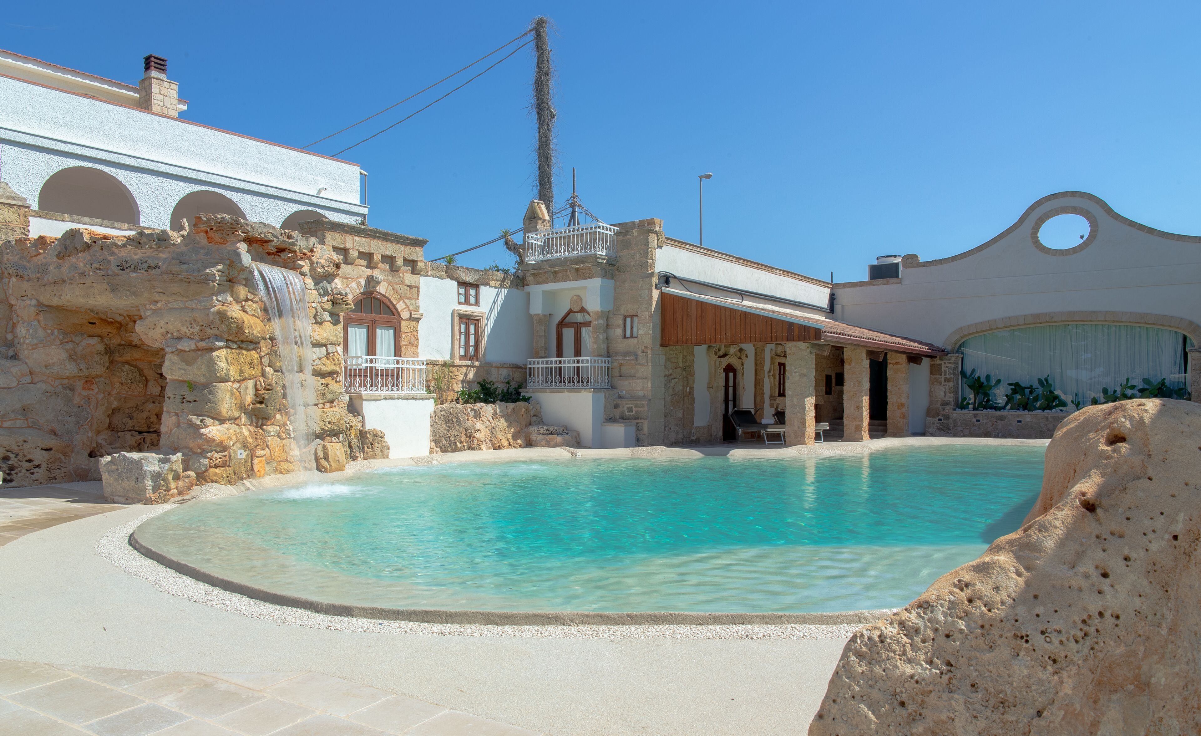 A pool area with a stone wall, a wooden deck, and a building with arched windows and a balcony.