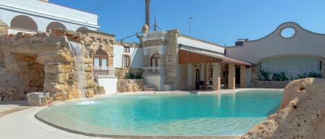 A pool area with a stone wall, a wooden deck, and a building with arched windows and a balcony.