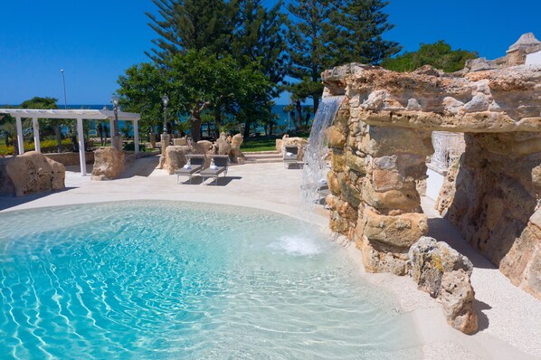 Une piscine à l’eau claire, une cascade décorative et une pergola blanche agrémentée de chaises longues.