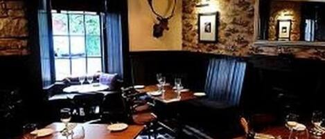 Un restaurant aux murs de pierre, orné d’une tête de cerf empaillée et doté d’une fenêtre avec des rideaux. Les tables sont dressées avec des assiettes, des verres et des couverts.