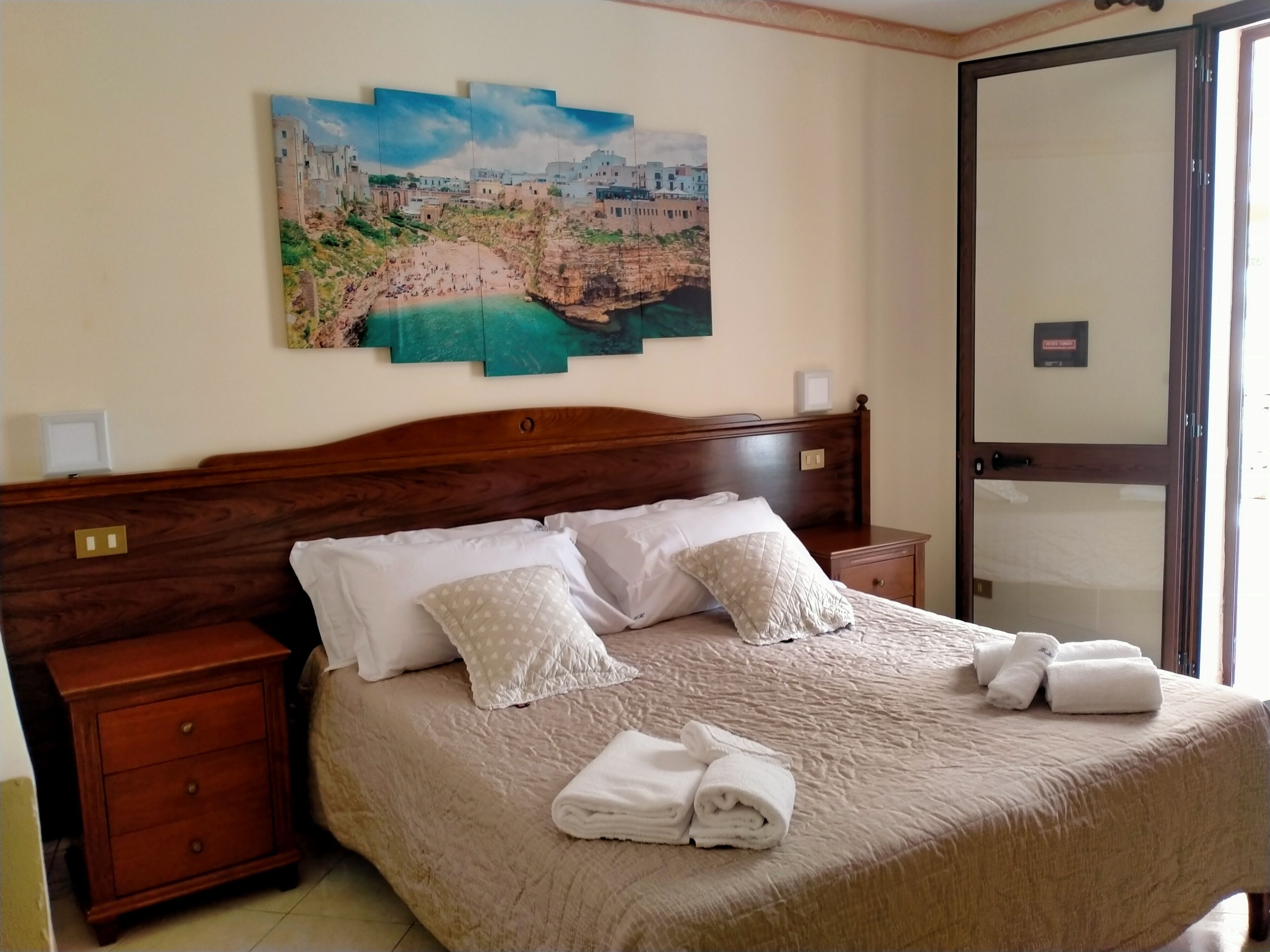 A neatly made bed with pillows and folded towels, a wooden nightstand, a wall-mounted painting of a coastal town, and a door with a sign.