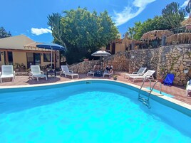 Piscina con sillones de descanso y sombrillas, rodeada por un muro de piedra y vegetación.