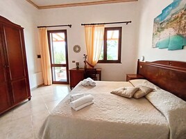 Un dormitorio con cama, dos ventanas con cortinas, un armario de madera, una mesita de noche con lámpara y un cuadro en la pared.