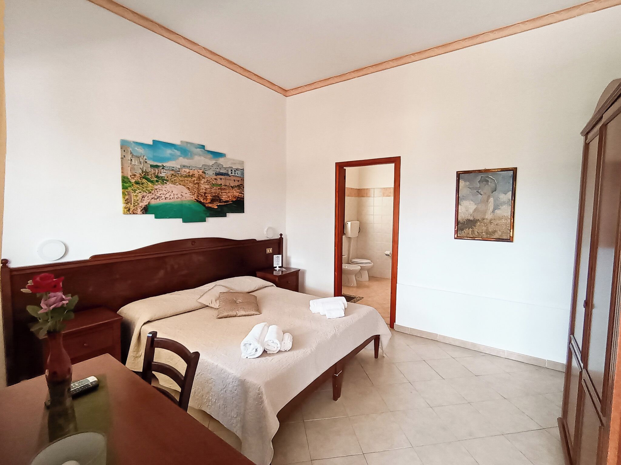 A bedroom with a bed, bedside table, a vase with flowers, a painting, and a bathroom visible through an open door.