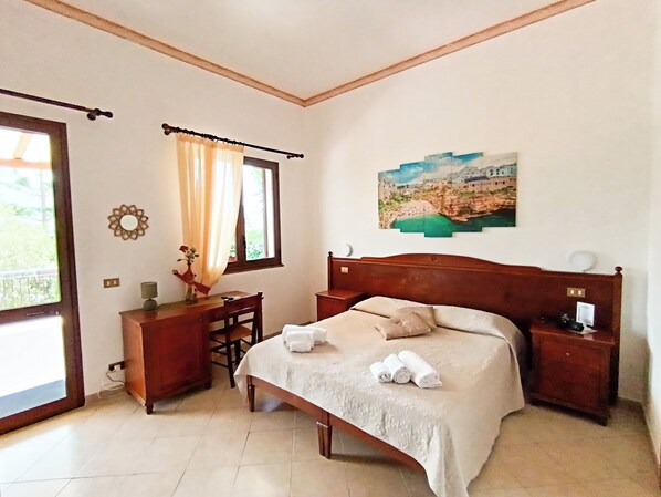 A bedroom with a bed, bedside tables, a desk, a chair, a window with curtains, a wall-mounted painting, and a decorative mirror. - HOTEL GROTTICELLI  (Castellammare del Golfo)