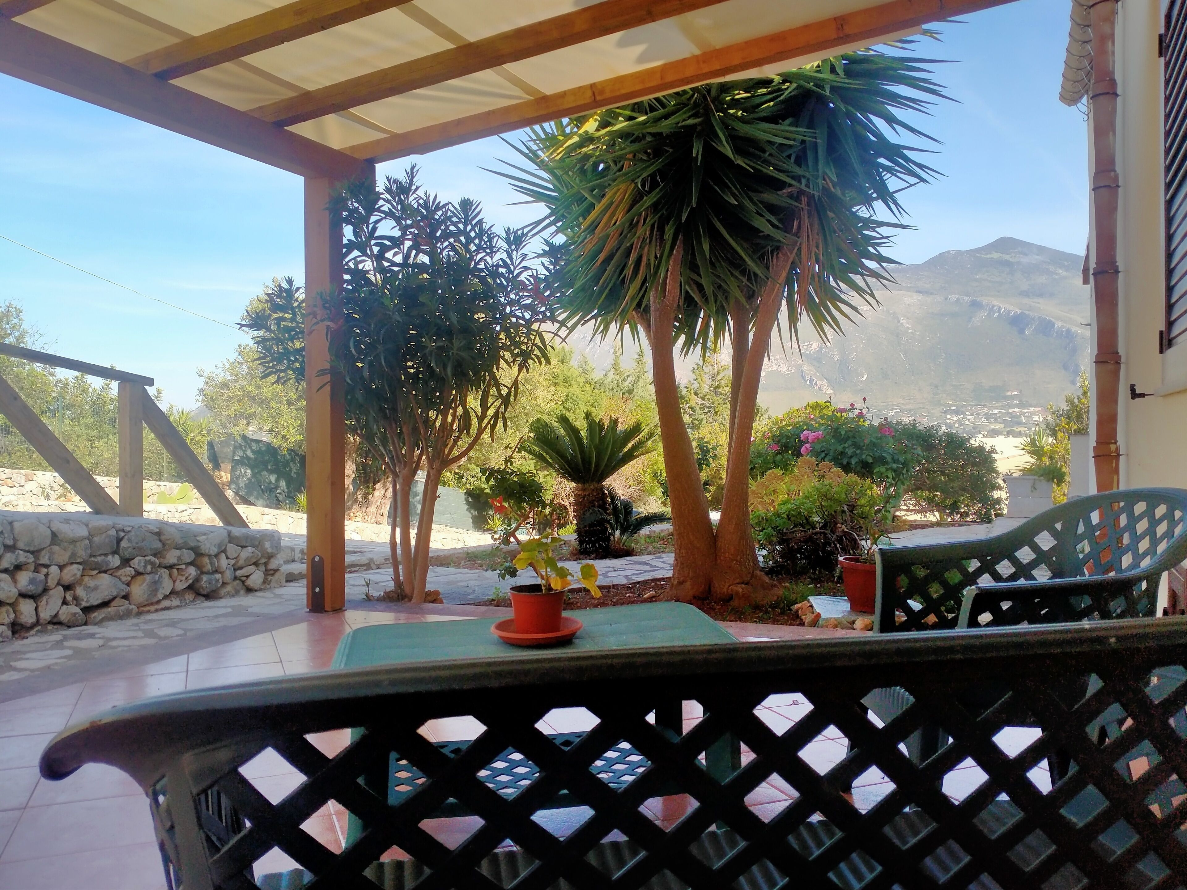 Um alpendre coberto com vista para uma montanha, com uma mesa, cadeiras e vários vasos de plantas.