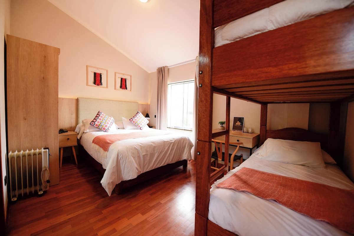 A bedroom with a wooden bunk bed, a single bed with patterned pillows, a bedside table, a nightstand, a radiator, and a window with curtains.