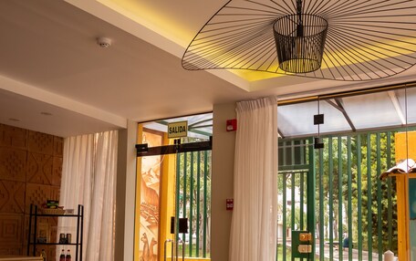 A modern hotel room with a round wooden table, two armchairs, a ceiling fan, and a view of greenery outside.