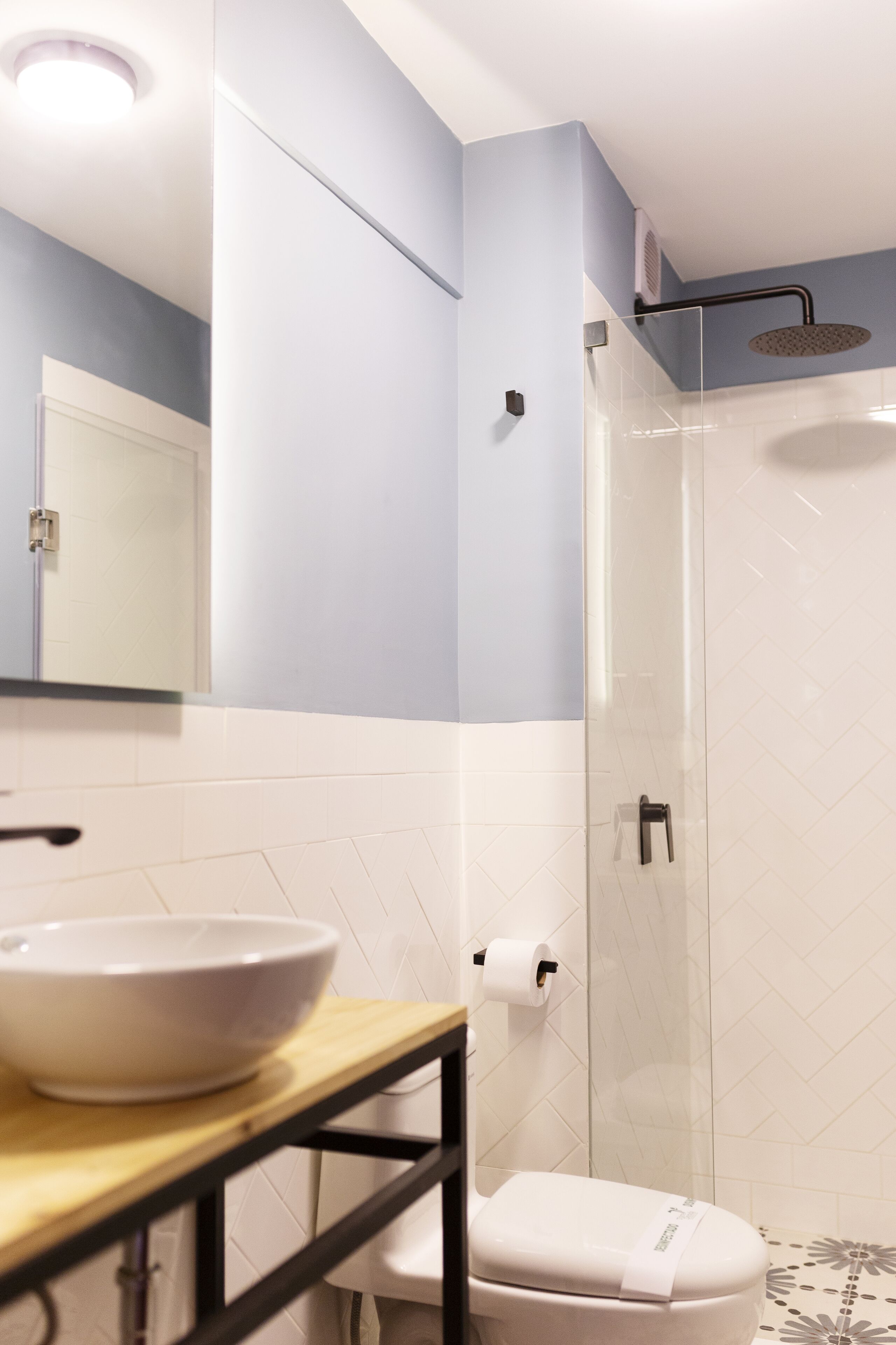 Un baño moderno con inodoro blanco, cabina de ducha con mampara de cristal, lavabo blanco sobre encimera de madera y un espejo sobre el lavabo.