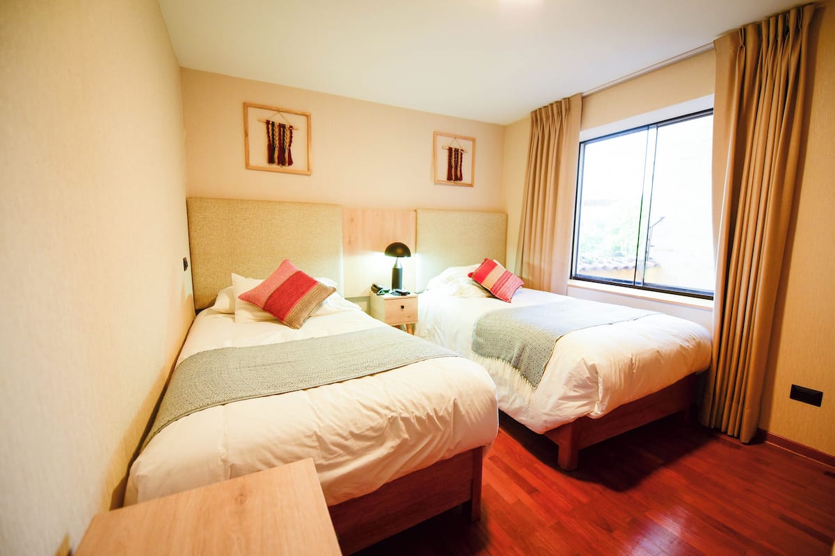 A hotel room with two beds, a nightstand, a lamp, and a window with curtains.