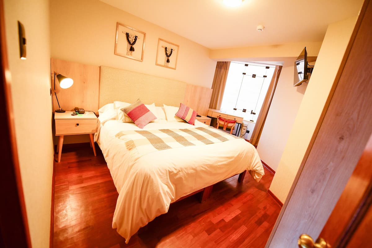 A bedroom with a wooden floor, a bed with a white comforter, a nightstand with a lamp, and a TV mounted on the wall.