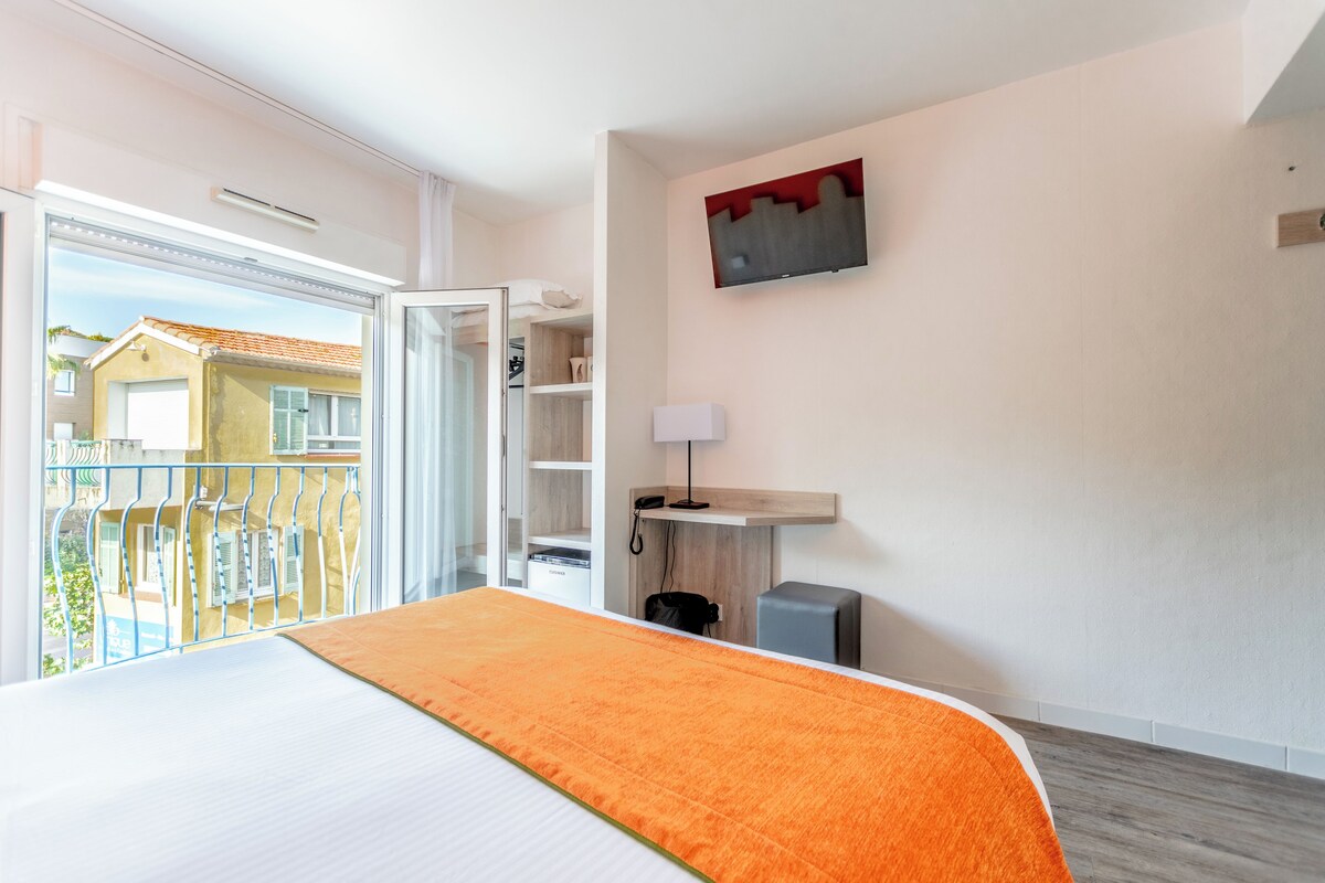 A hotel room with a bed, a balcony, a wooden floor, a wall-mounted TV, and a small desk with a lamp.