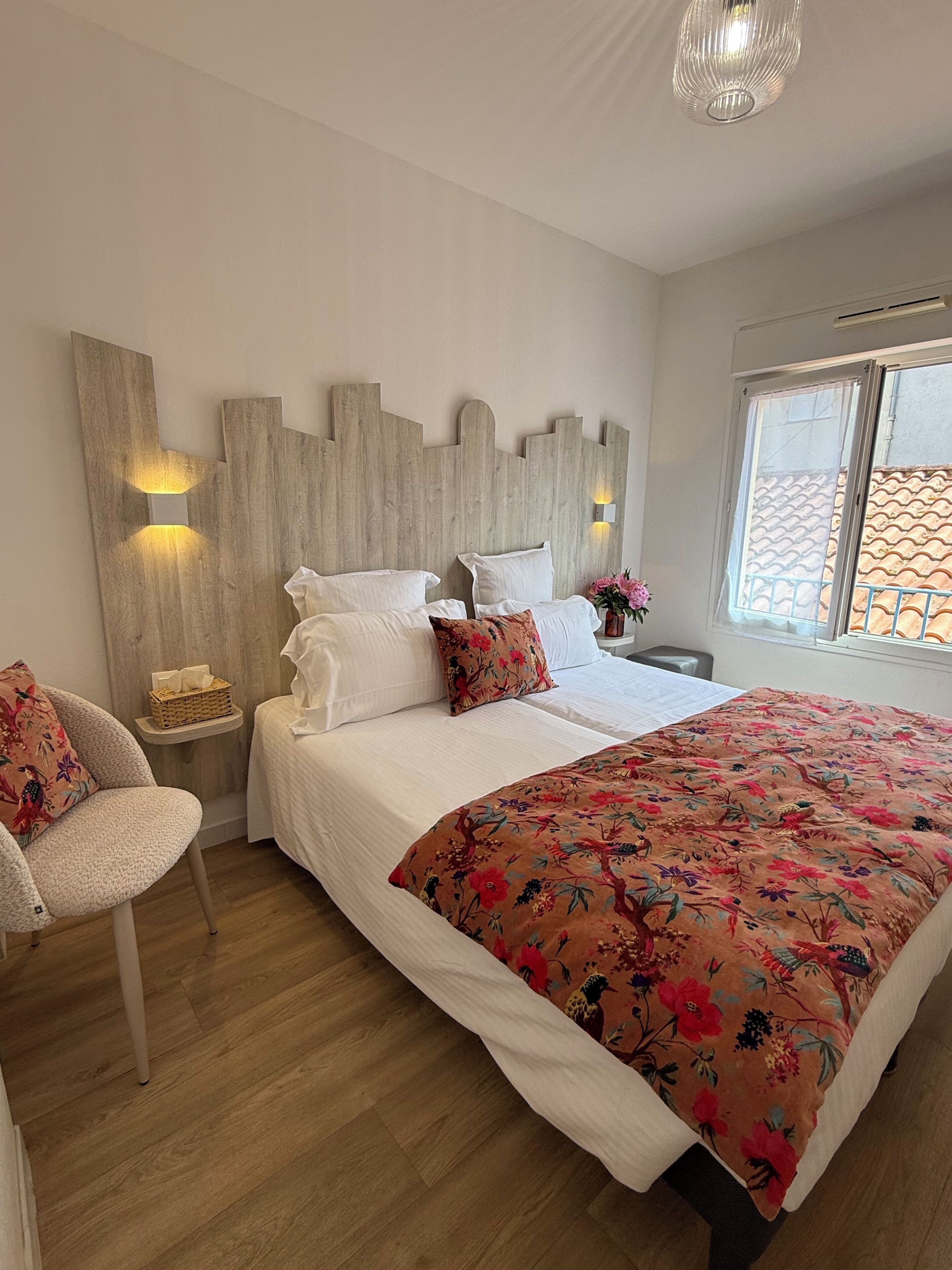 A bedroom with a wooden headboard, a bed with white linens, a floral bedspread, a chair, a nightstand with a lamp, and a window with a view.