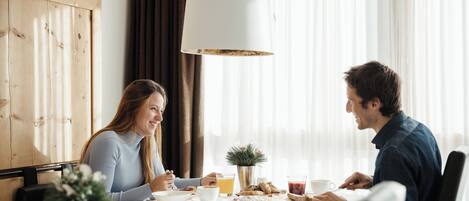 A dining area with a table set for two, featuring a white tablecloth, a potted plant, and a variety of breakfast items.