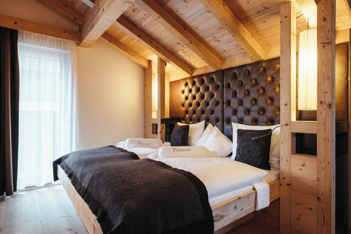 A wooden-framed bedroom with a tufted headboard, a large bed with white linens, and a window with sheer curtains.