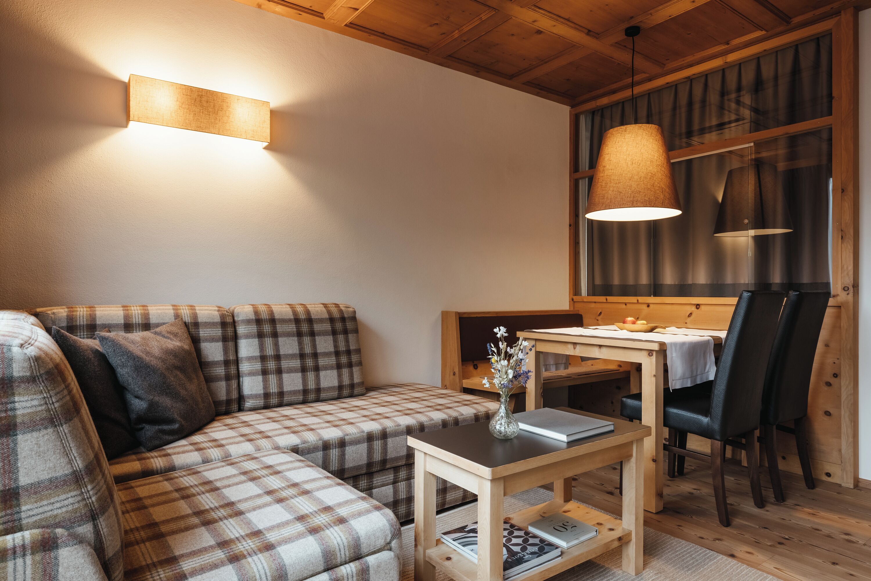 A cozy living room with a plaid sofa, a wooden table with a vase, and a dining area with chairs.