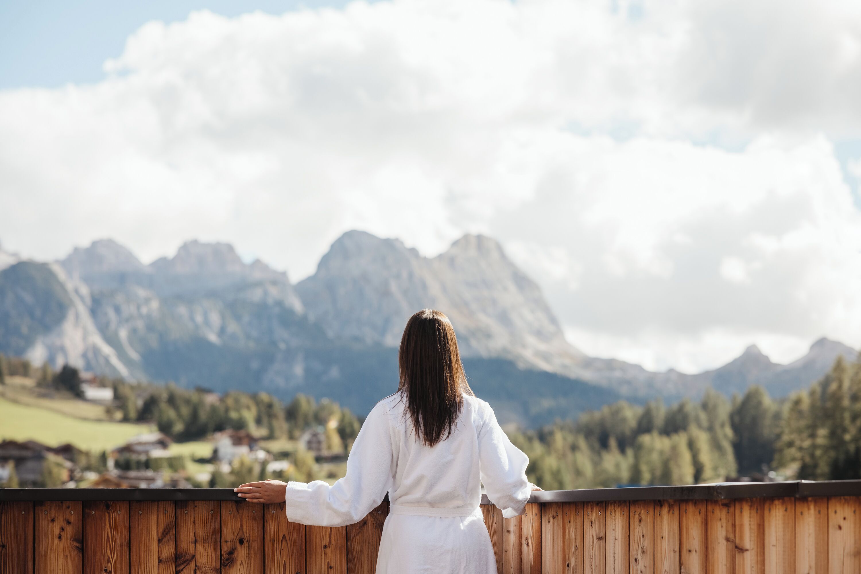 A person standing on a wooden balcony overlooking a mountainous landscape with houses in the valley below.