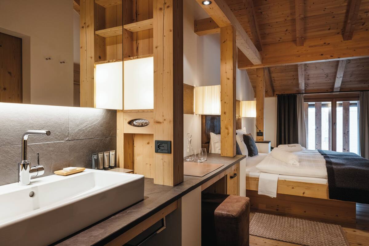 A modern bathroom with a large sink, wooden beams, and a bed with a dark blanket.