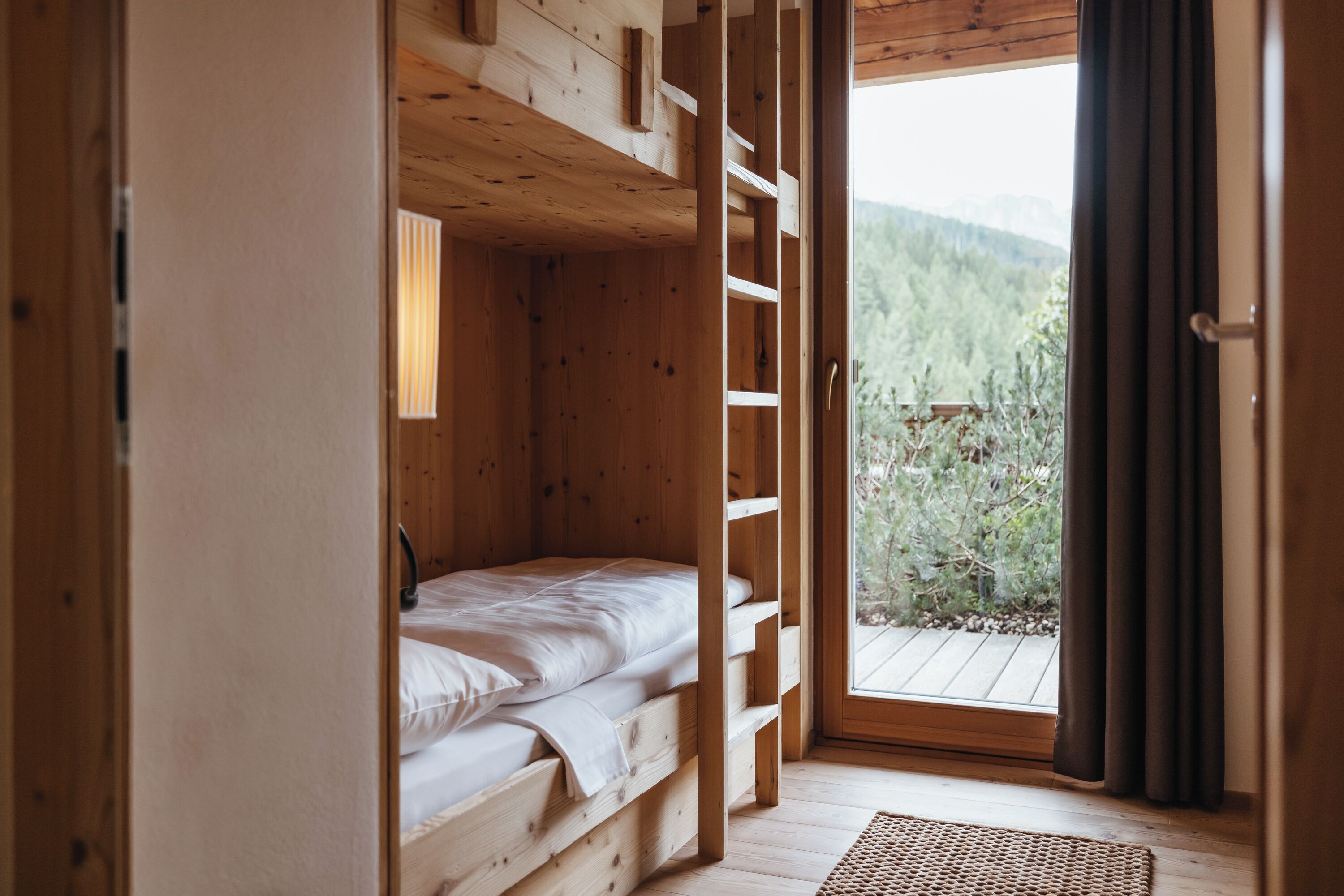 A wooden cabin room with a bunk bed, a window with a view of a forest, and a door leading outside.