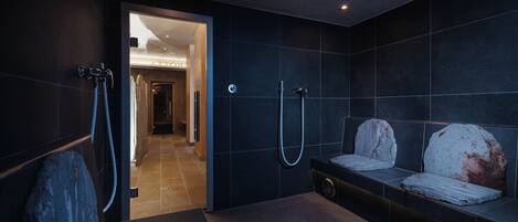 A modern bathroom with dark tiles, a shower area, and a bench with towels.