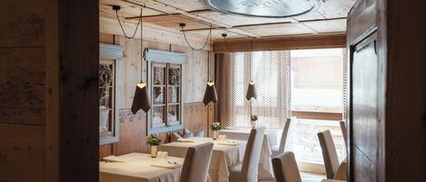 A rustic dining area with wooden walls and ceiling, featuring tables set for dining, pendant lights, and a large window with patterned curtains.
