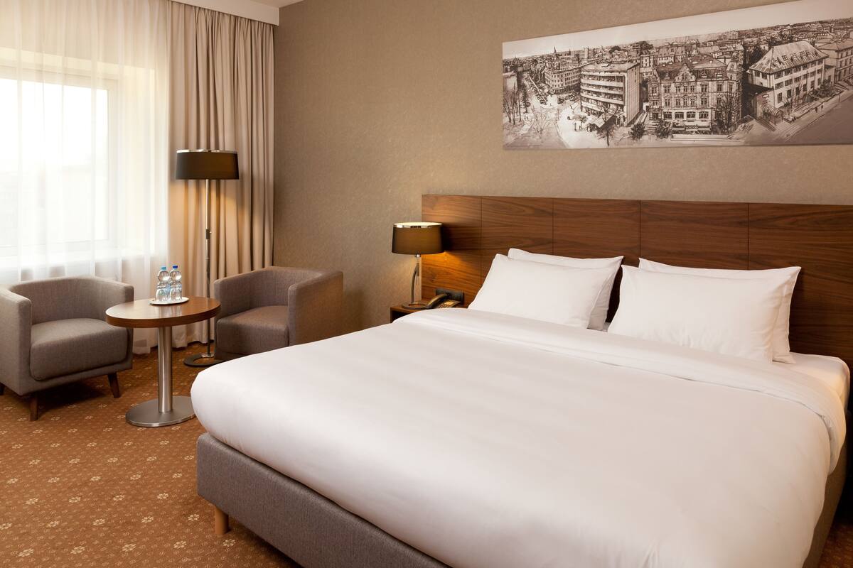 Una habitación de hotel con una cama grande, dos sillones, una mesita redonda y una lámpara.