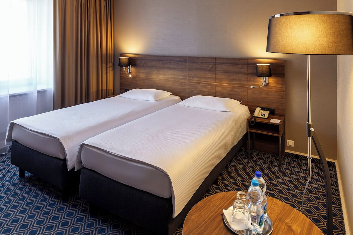Habitación de hotel con dos camas, una mesita de noche con lámpara, teléfono y un recipiente con botellas de agua.