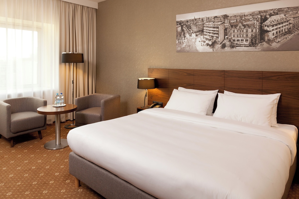 A hotel room with a large bed, two armchairs, a small round table, and a lamp.