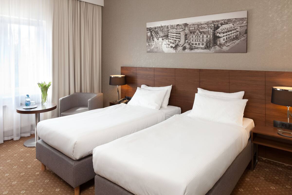 Habitación de hotel con dos camas individuales, una mesita con lámpara, una silla y un ventanal con cortinas traslúcidas.