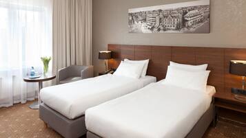 A hotel room with two single beds, a small table with a lamp, a chair, and a large window with sheer curtains.