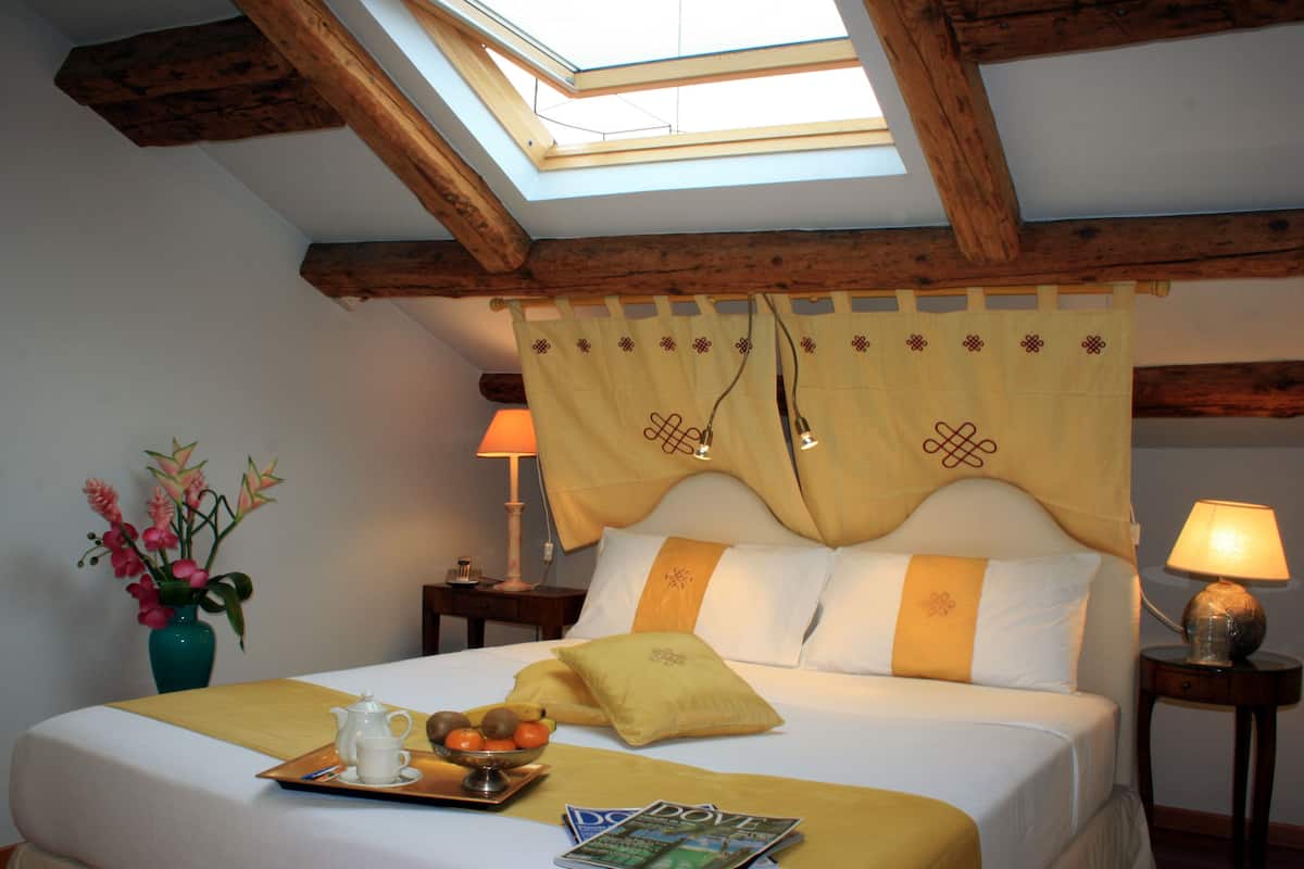 Habitación de hotel con cama, mesita de noche, lámpara, un florero con flores y una bandeja con tetera y fruta.