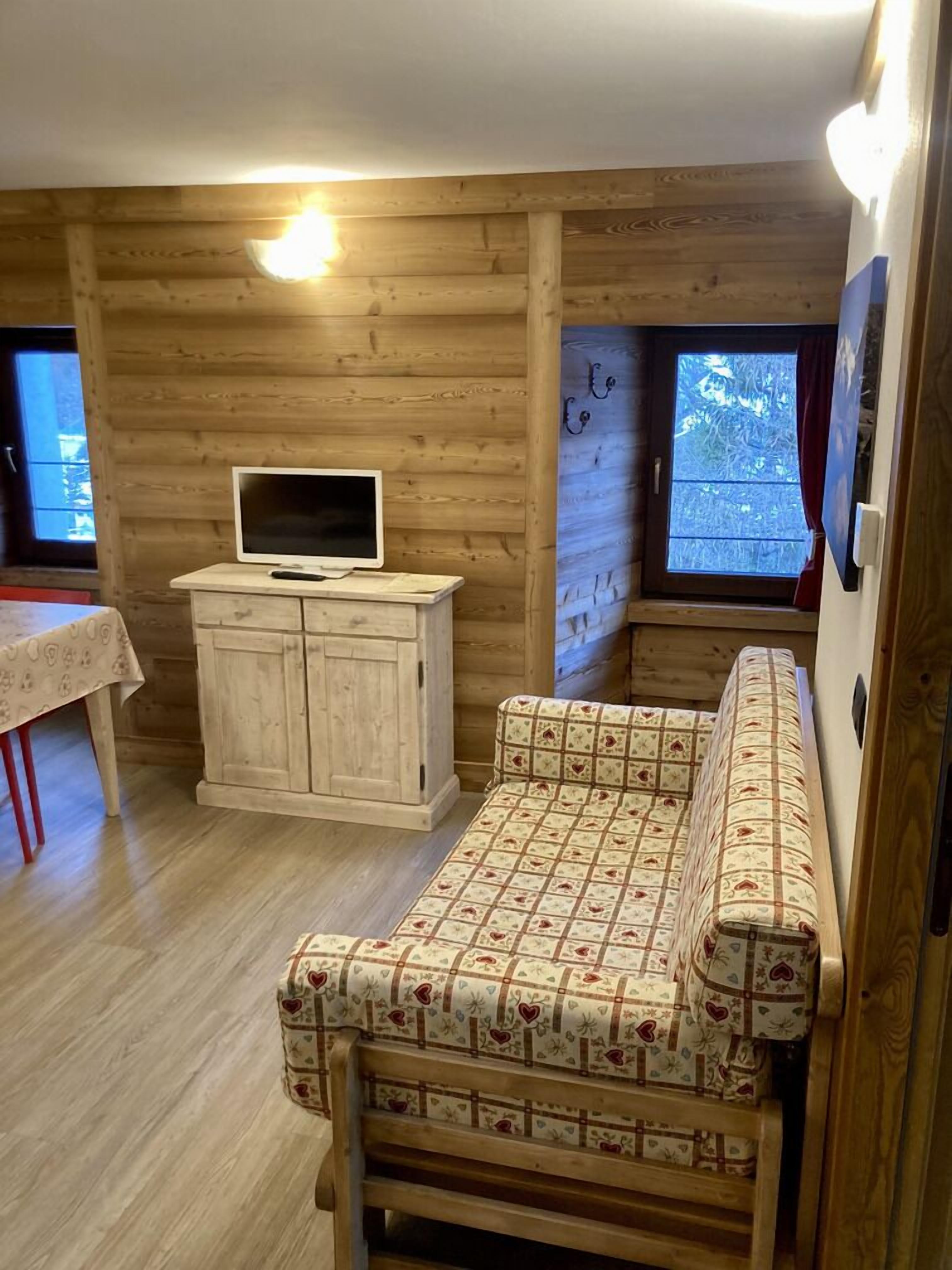 A cozy cabin interior with a wooden wall, a small white cabinet with a TV, a bench with a patterned cushion, and a window with a view of trees.